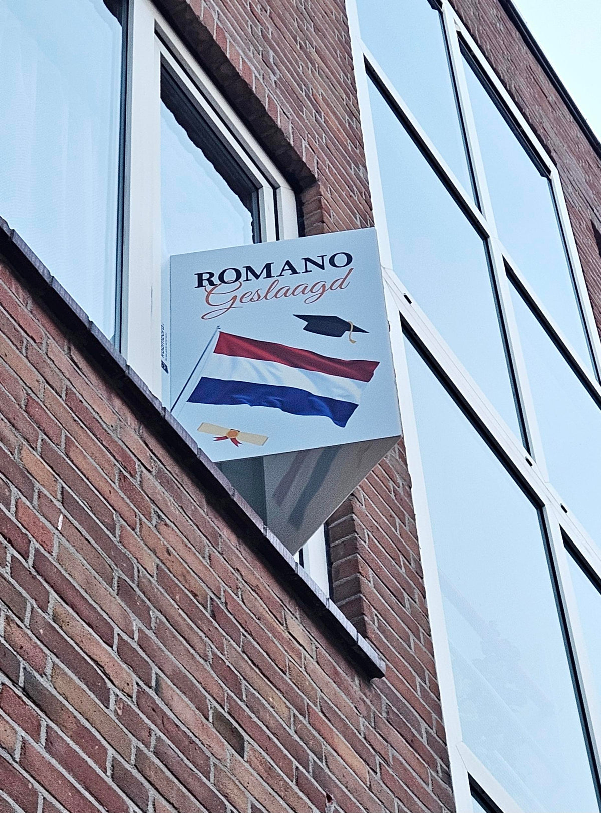 Geslaagd-raambord met Nederlandse vlag aan een raam in een bakstenen gevel